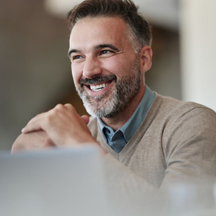 A smiling man with short brown hair and a salt-and-pepper beard, wearing a light brown sweater over a blue collared shirt, sits at a table with his hands clasped, looking slightly to the side.