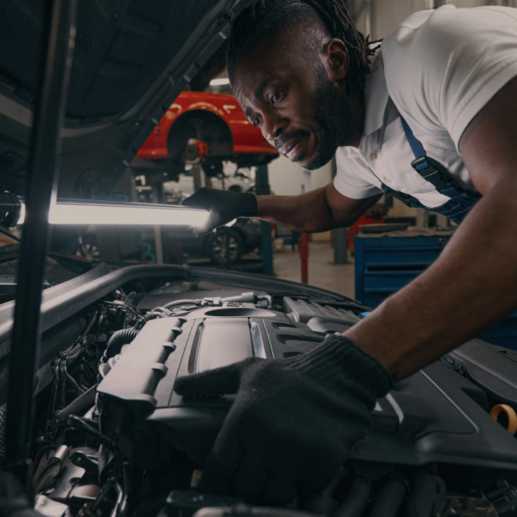 A mechanic wearing gloves and overalls examines a car engine under the hood in a garage, using a bright work light for visibility. Another red car is visible lifted in the background.