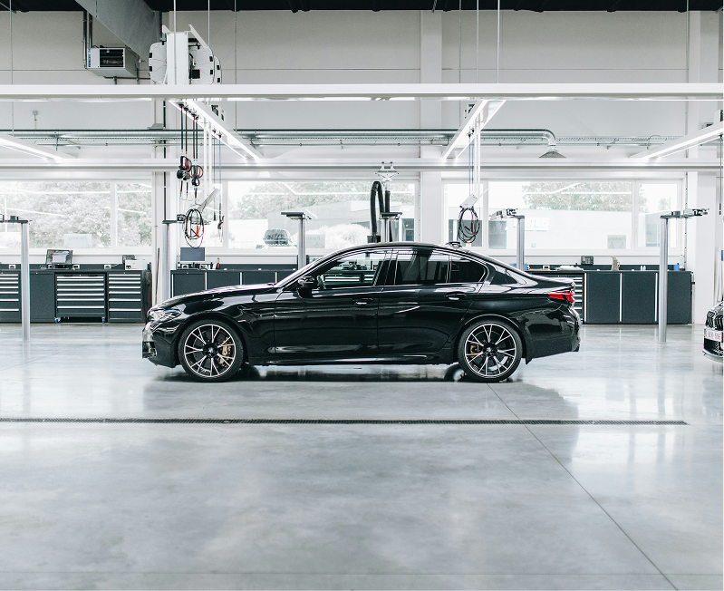 A sleek black luxury sedan is parked inside a modern, spacious automotive workshop with large windows, workbenches, and overhead lights.